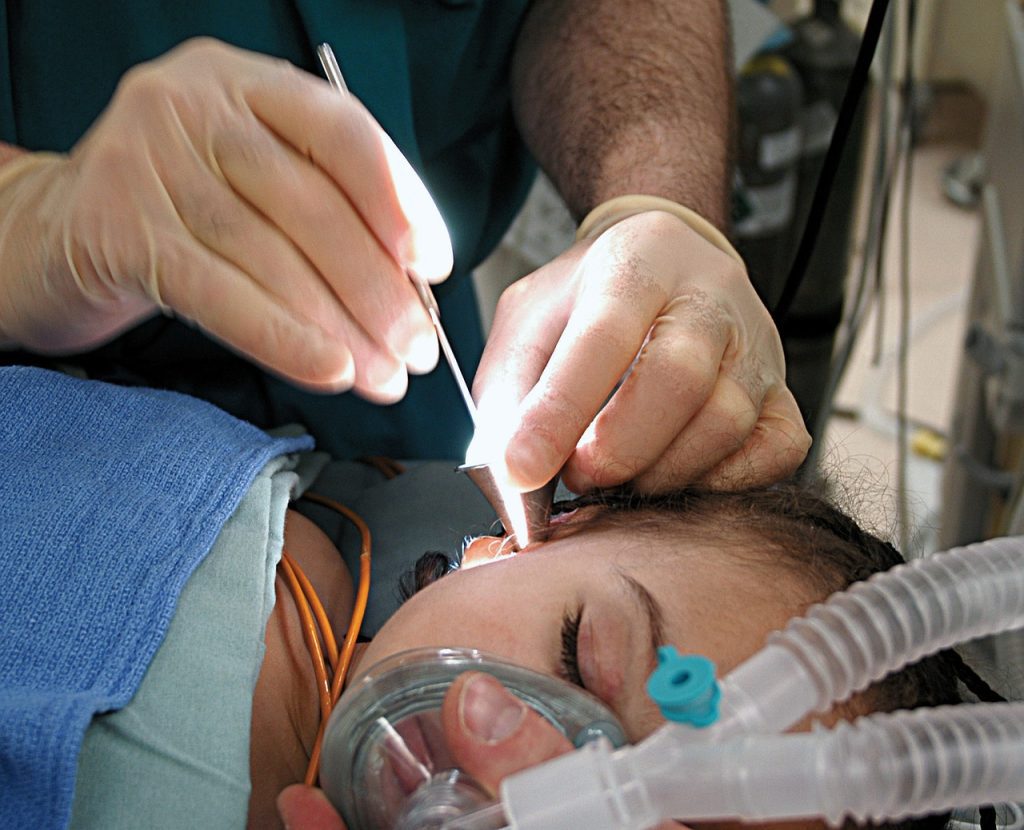 A doctor performs a medical procedure on a patient using a tool with a bright light, while the patient is under anesthesia with an oxygen mask. The grownup patient's family waits anxiously in the waiting