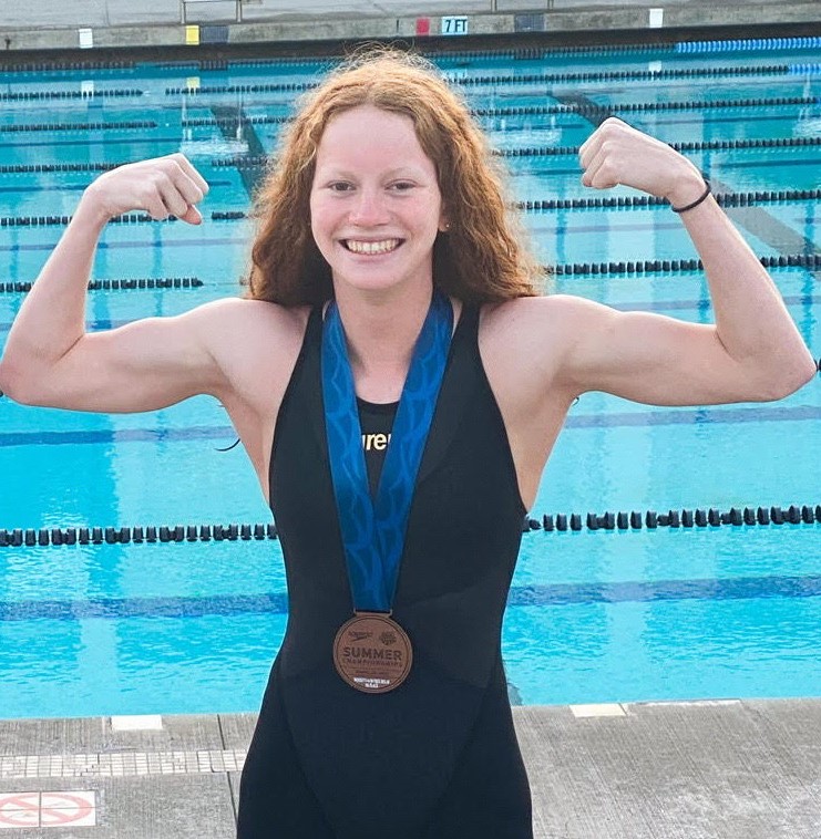 A smiling female swimmer with a hearing disability, wearing a medal stands by a pool, flexing her muscles in a victory pose.