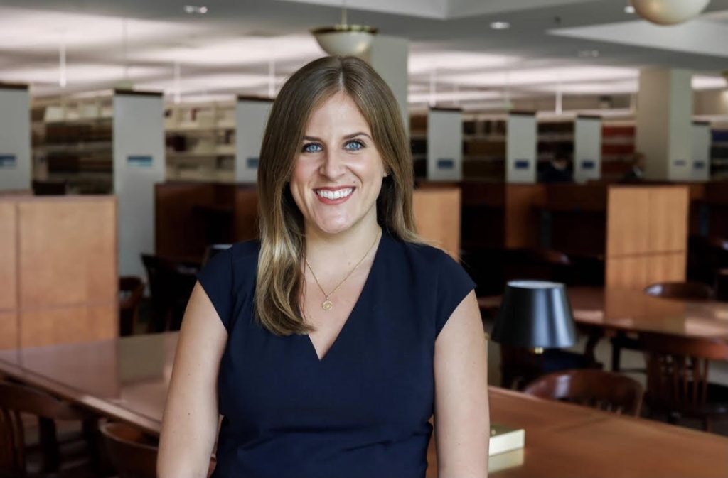 A smiling grownup woman with long blonde hair, wearing a navy blue top, standing in a library with bookshelves and reading desks in the background.