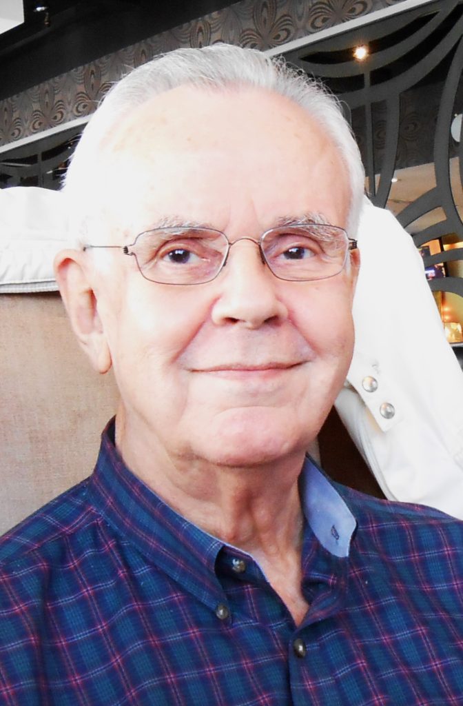 Elderly man with glasses smiling in a plaid shirt, indoors with a family photo patterned background.