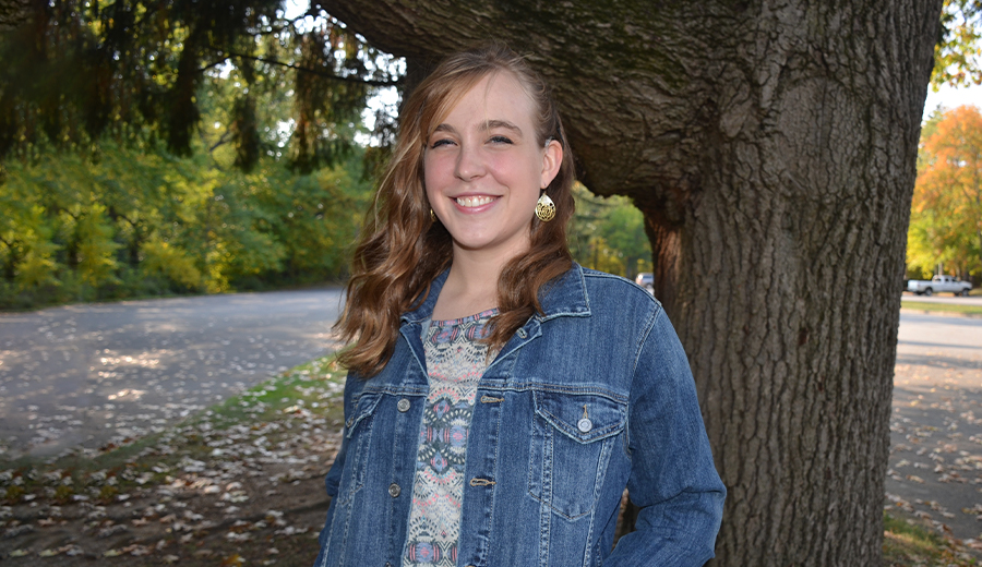 Young woman in a denim jacket smiling, standing by a tree in a park during autumn with her child.