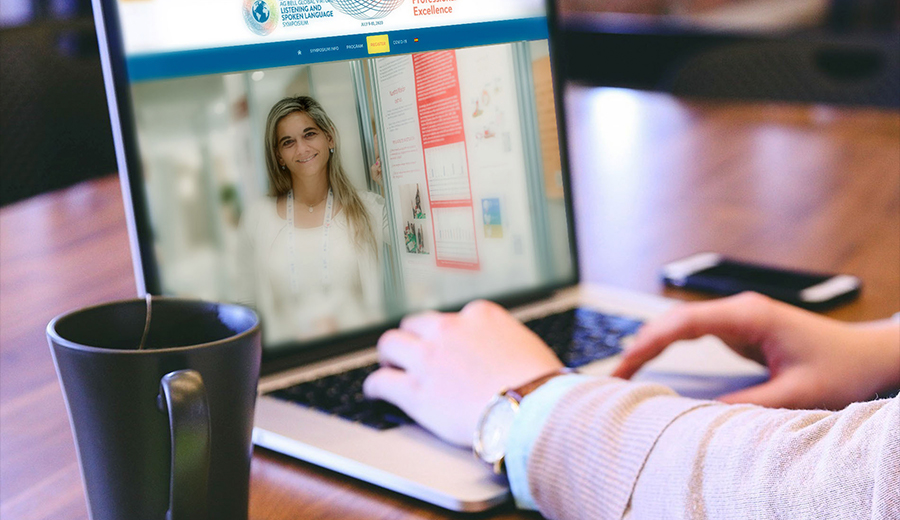 A person browsing a laptop with a smiling woman on the screen, featuring an educational website, alongside a coffee mug on a wooden desk. In the background, you can hear children playing.