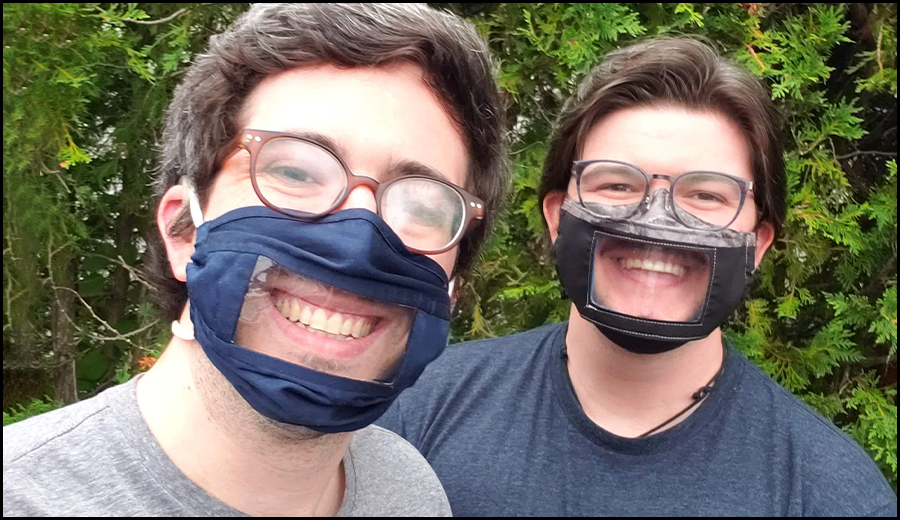 Two men, possibly family members, smiling and wearing face masks with clear panels over the mouth area, standing outdoors with greenery in the background.