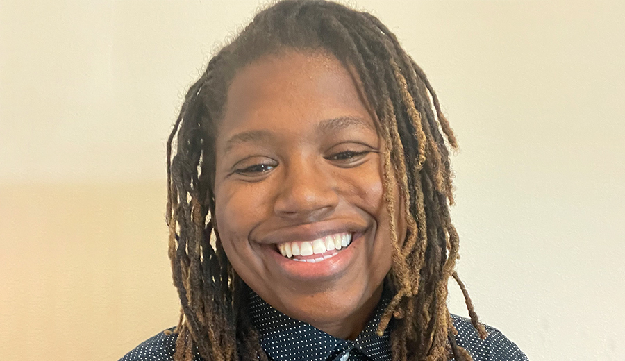 A grownup with a broad smile, wearing a polka-dot shirt, and sporting twisted dreadlocks, standing against a beige wall.