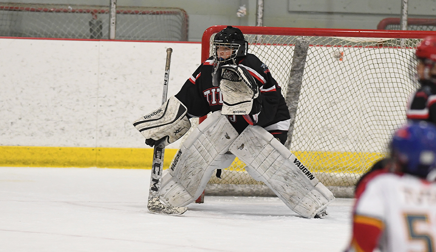 A hockey goalie in black and red gear prepares to block a shot on ice, with assistance from players visible in the background.