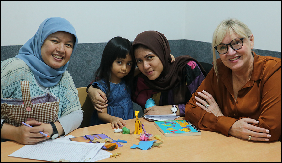 Four women, one child with a disability, and diverse backgrounds, sitting around a table with educational toys and papers, smiling at the camera in a classroom.