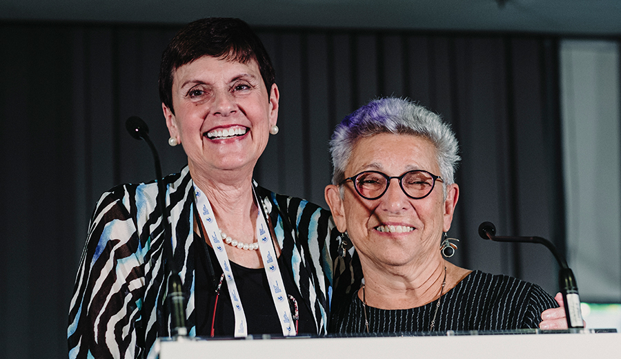 Two smiling women, one with short black hair and a striped top, the other with gray hair and glasses, standing together behind a podium to provide assistance.