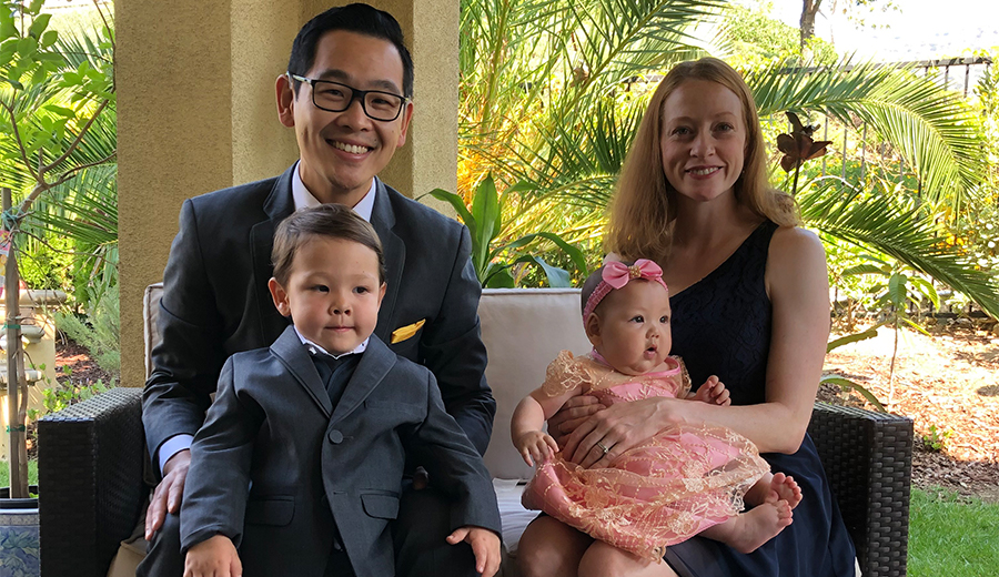 A family of four, including a smiling grownup couple, a young boy, and a baby girl, dressed formally and sitting together on a patio.