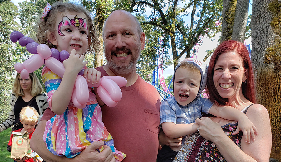 A family of four at an outdoor event, with the parents holding their children who have face paint and balloon animals, seeking assistance from a coordinator.