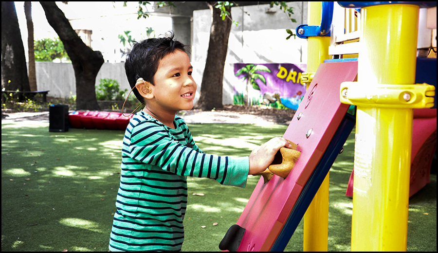 A young boy with a hearing disability in a striped shirt smiling while playing on colorful playground equipment outdoors.