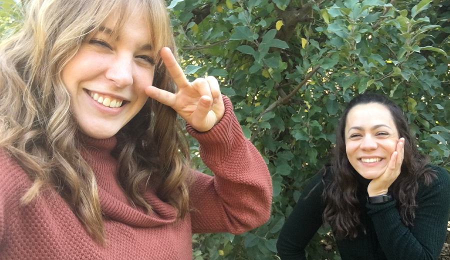 Two women smiling outdoors, one making a peace sign, standing near green foliage with family.