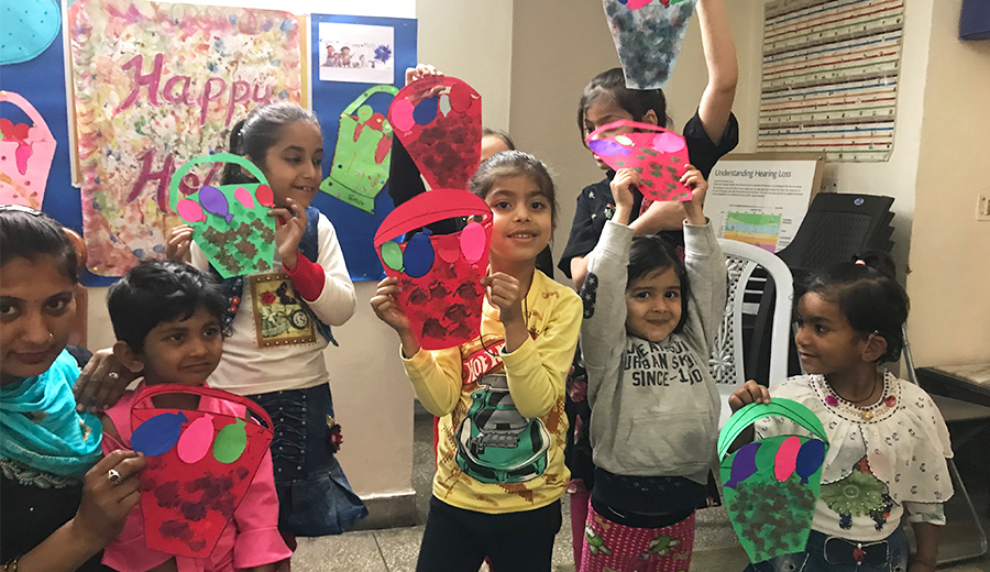 Children showing their colorful paper craft projects in a classroom with a "happy holidays" poster in the background, alongside family members who have come to hear about their work.
