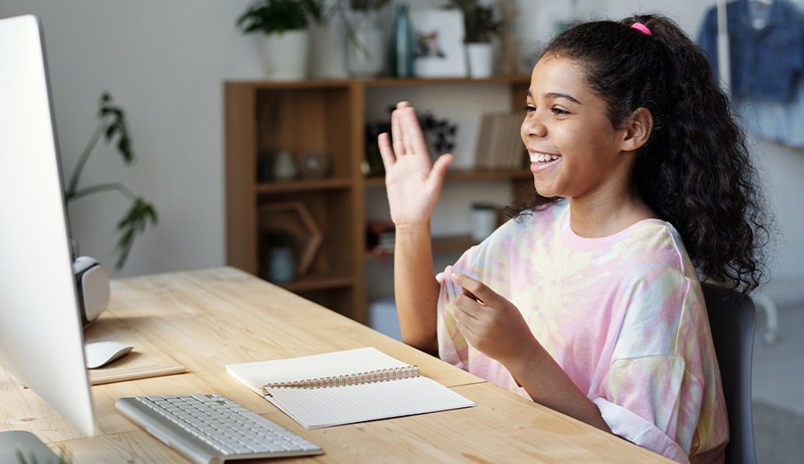 A young child smiles and waves at a computer screen, sitting at a desk with a notebook and keyboard in an office setting.