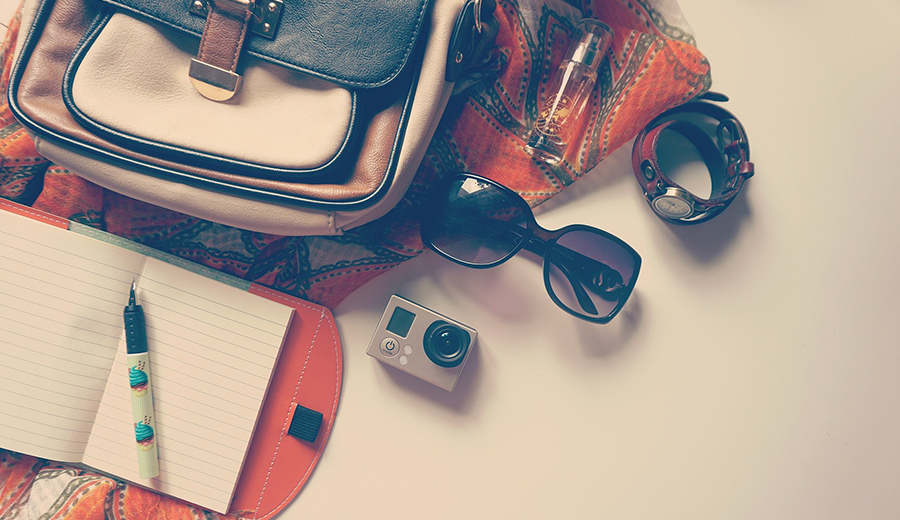 Flat lay of travel essentials including a notebook, pen, sunglasses, camera, wristwatch, scarf, hearing aid, and satchel on a light surface.