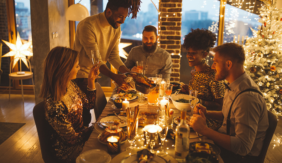 Group of friends enjoying a Christmas dinner together in a warmly lit room decorated with a Christmas tree and lights, discussing how to better assist those with disabilities.
