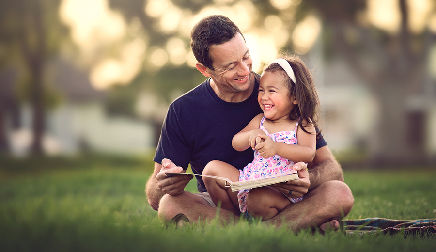 A father and his young daughter, who has a disability, sitting on grass, laughing together while reading a book at sunset.