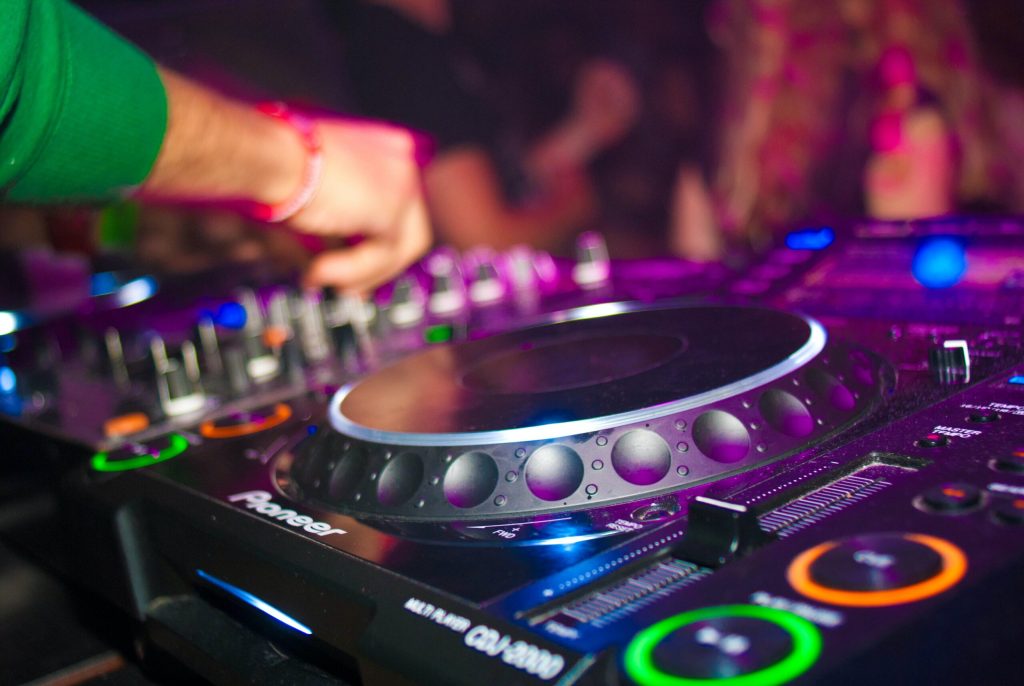 A DJ's hand adjusting knobs on a Pioneer DJ mixer at a vibrant nightclub event, with colorful lights in the background as family members gather to hear the set.