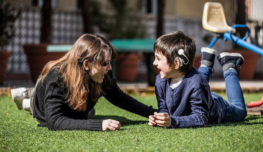 A woman and a young boy with a disability lying on grass, facing each other and laughing, with playground equipment in the background.