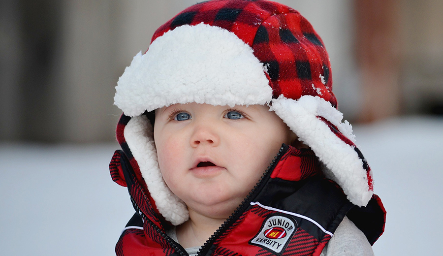 A child with blue eyes, wearing a red and black plaid hat and a jacket, looks to the side in a snowy setting.