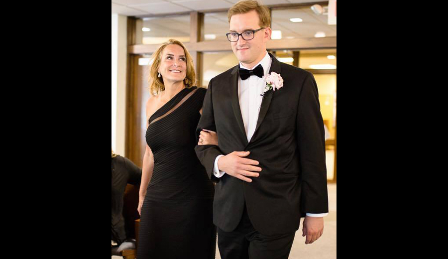 A man in a black tuxedo and glasses, supporting a woman with a mobility disability in an elegant black dress, walks arm-in-arm at a formal event.