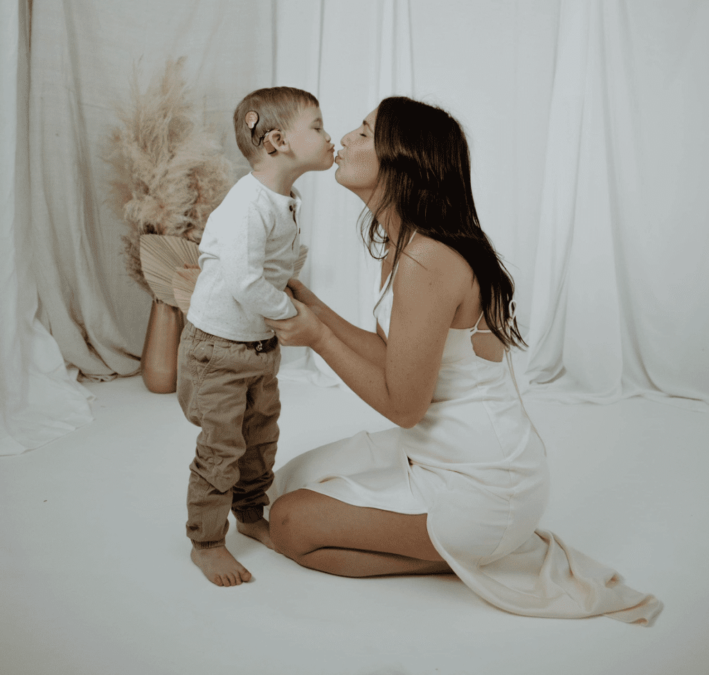 A young boy in a white shirt and beige pants, with a hearing aid, kissing a woman in a white dress, in a studio with a white backdrop.