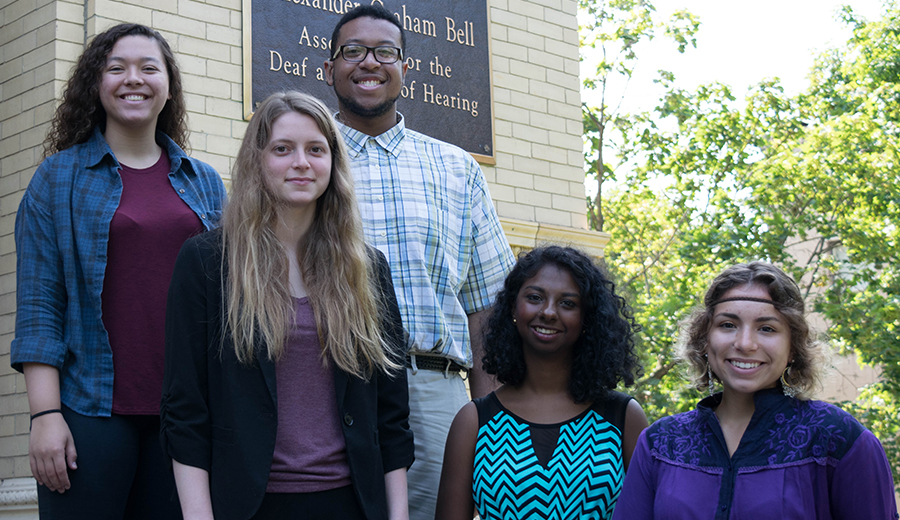 Five diverse young adults smiling in front of a building with a sign reading "Alexander Graham Bell Association for the deaf and hard of hearing, offering assistance to families.