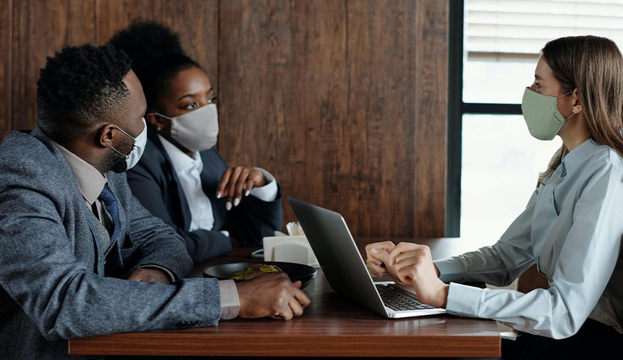 Three professionals wearing masks sit at a desk during a meeting, one of them using a laptop to assist with documentation.