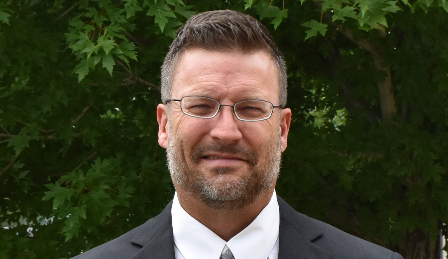 A man with short hair and a beard, wearing glasses and a dark suit, smiles gently in front of a tree with lush green leaves as he listens to the laughter of a child nearby.