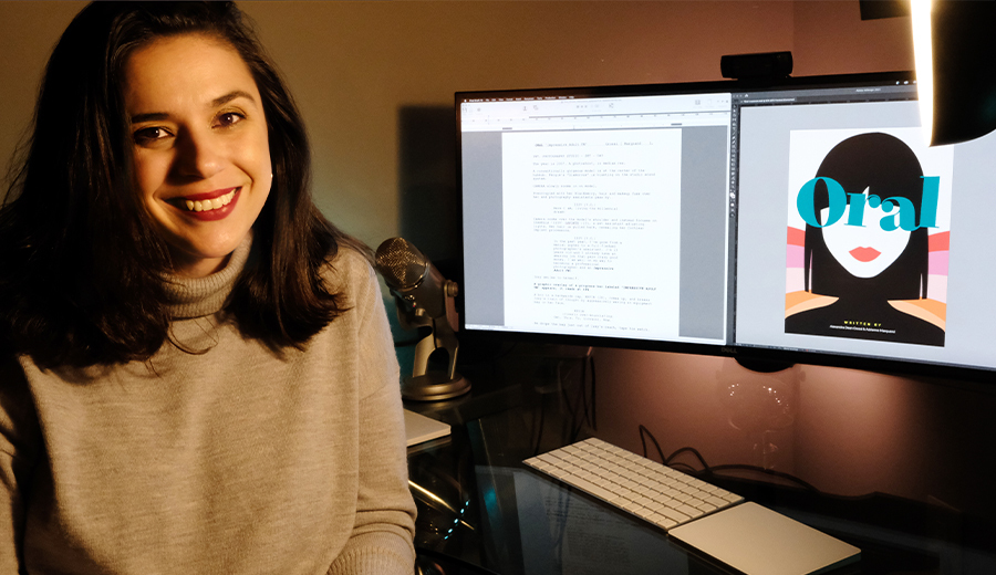 Woman smiling at camera in a home office, with two computer screens displaying text and graphic design work related to disability awareness.