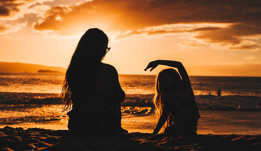 Two silhouetted family members on a beach at sunset, one standing and the other stretching an arm overhead, with the ocean in the background.