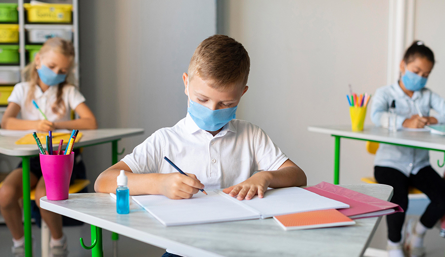 A young child wearing a face mask writes in a notebook at his desk in a classroom with other masked students in the background.
