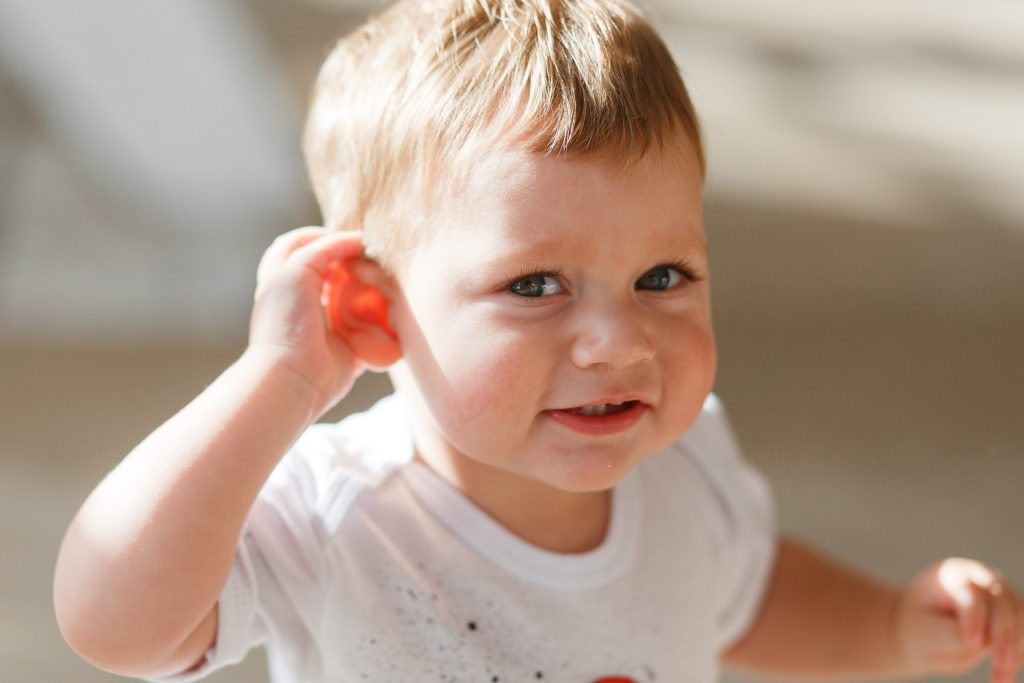 Toddler with a smile tugging on ear, sunlight illuminating hair, wearing a white shirt, needing assistance.