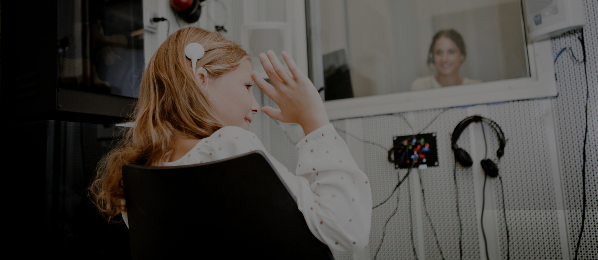 Young girl with a headset sitting in front of a computer, waving at the screen where a smiling woman appears, in a home office setting.